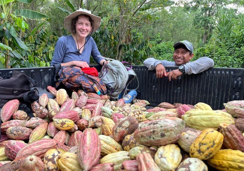 A man and a women lean on the bed of a pick-up truck filled with cacao pods that are yellow, pink, and red in color.